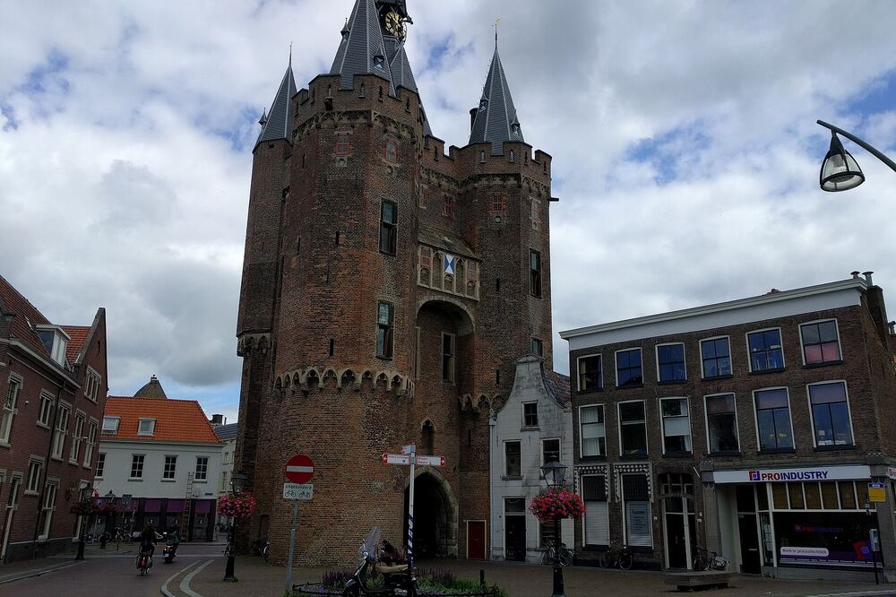 De middeleeuwse Sassenpoort in Zwolle met vier torenspitsen, gezien vanaf het plein met winkelpanden, hangende bloembakken en fietsers ervoor
