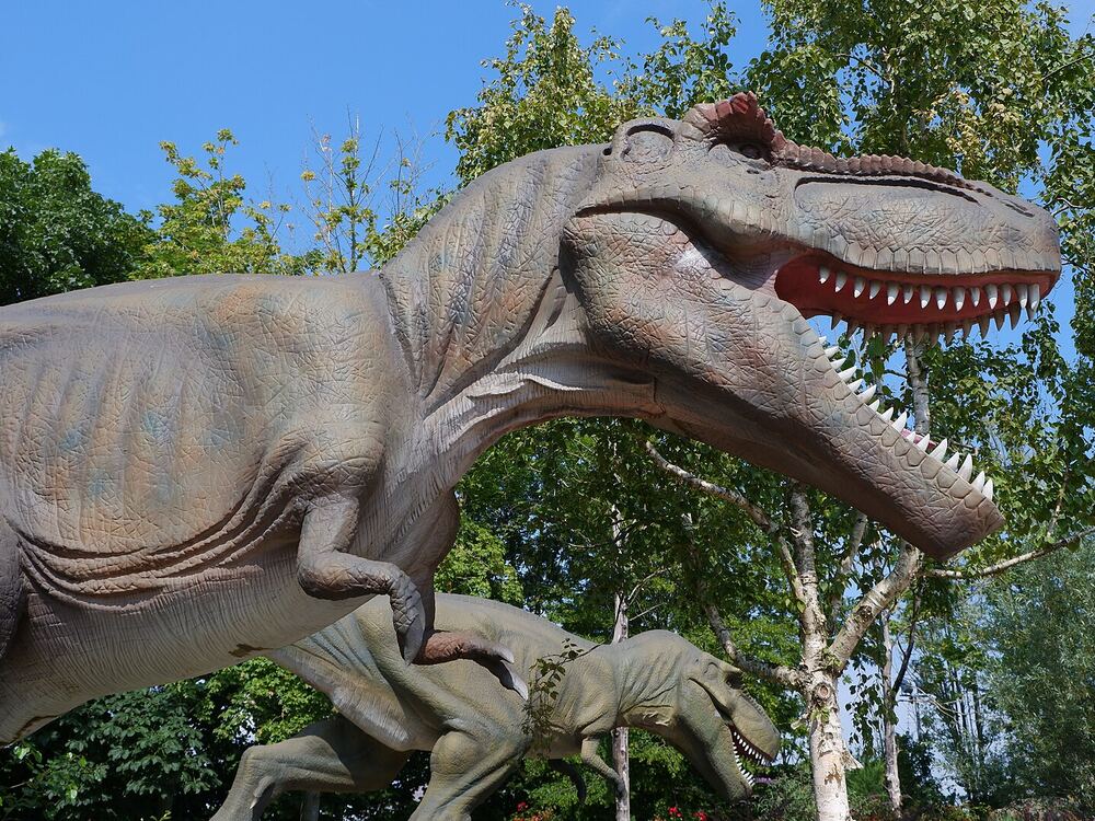 Levensgroot beeld van een tyrannosaurus met open bek tussen groene bomen in Dinoland Zwolle, met een kleinere dinosaurus ernaast
